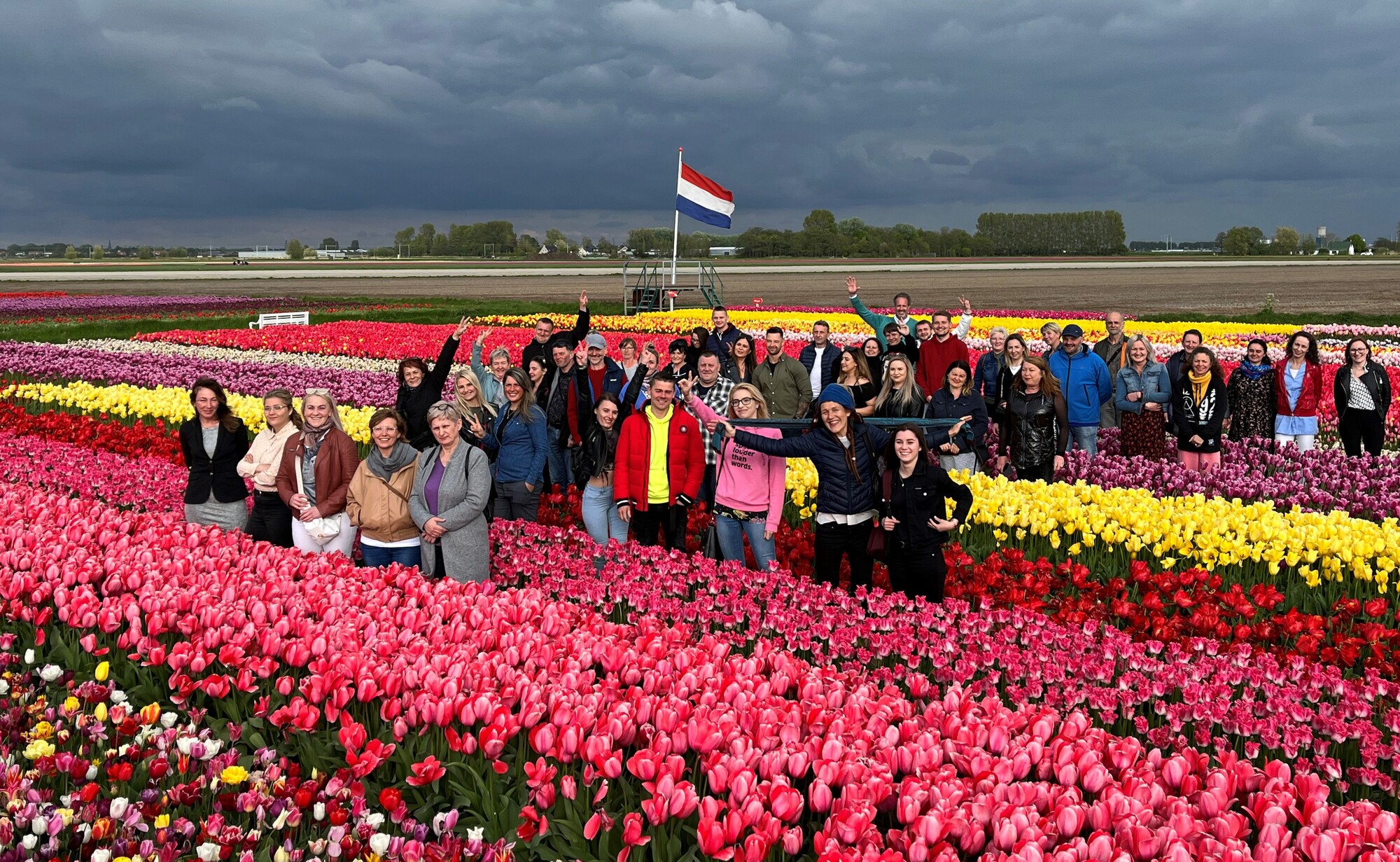 Vrolijke groep geniet van een tulpenveld, met de Nederlandse vlag op de achtergrond. Ondanks het slechte weer, tulpen tuinen Nederland, Amsterdamse tulpenrondleiding. Kleurrijke bloemenvelden